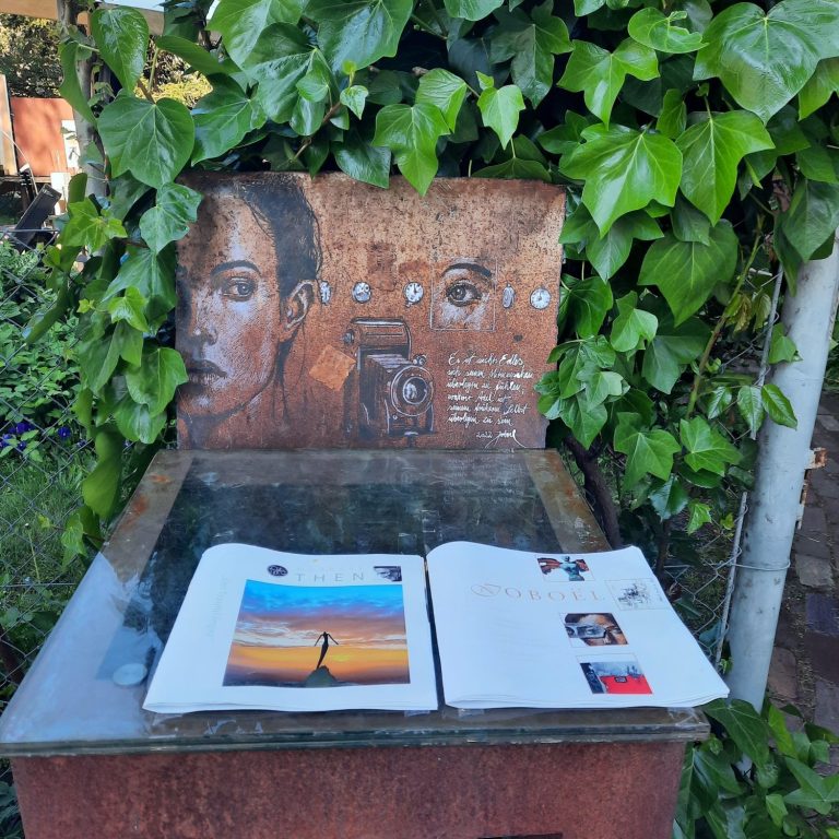 Ein Holztisch mit einer Künstlermarke und offenen Zeitschriften, umgeben von Pflanzen.