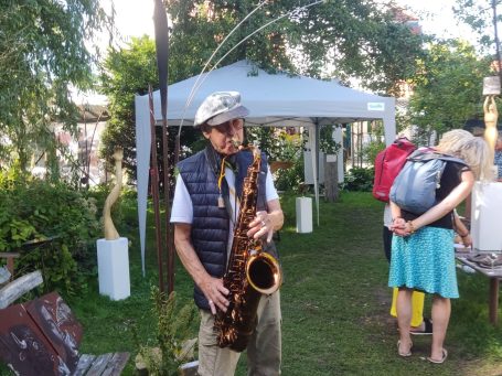 Ein Mann spielt Saxophon in einer Gartenumgebung mit Besuchern im Hintergrund.