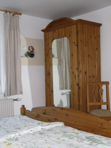 Holzschrank mit Spiegel und Stuhl, neben einem Bett in einem hellen Raum.