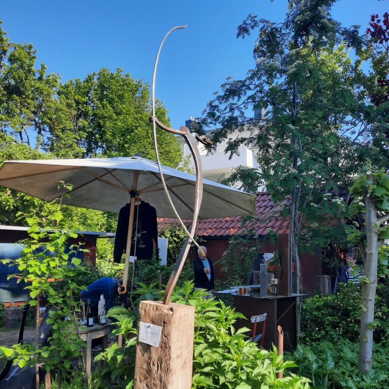 Skulptur aus Holz vor einem schattigen Sitzbereich mit Bäumen und Sonnenschirm.