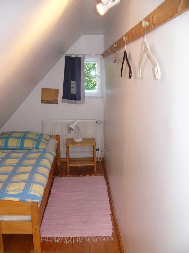 Kleines Schlafzimmer mit Bett, Teppich, Fenster und Kleiderbügel an der Wand.